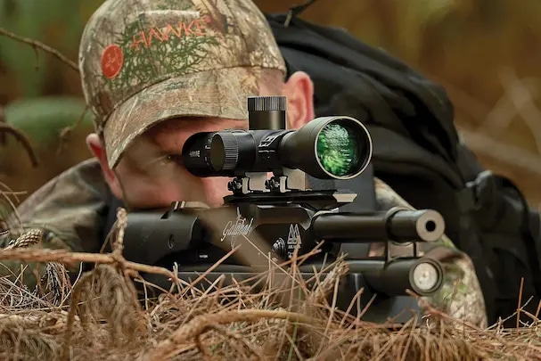 Hunter aiming through a Hawke rifle scope while lying in natural woodland cover, highlighting the scope’s clarity and precision in outdoor conditions.